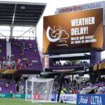 Soccer stadium with purple seats and a large LED screen showing 'Weather Delay! Match will restart as soon as possible' and a goal in the foreground.
